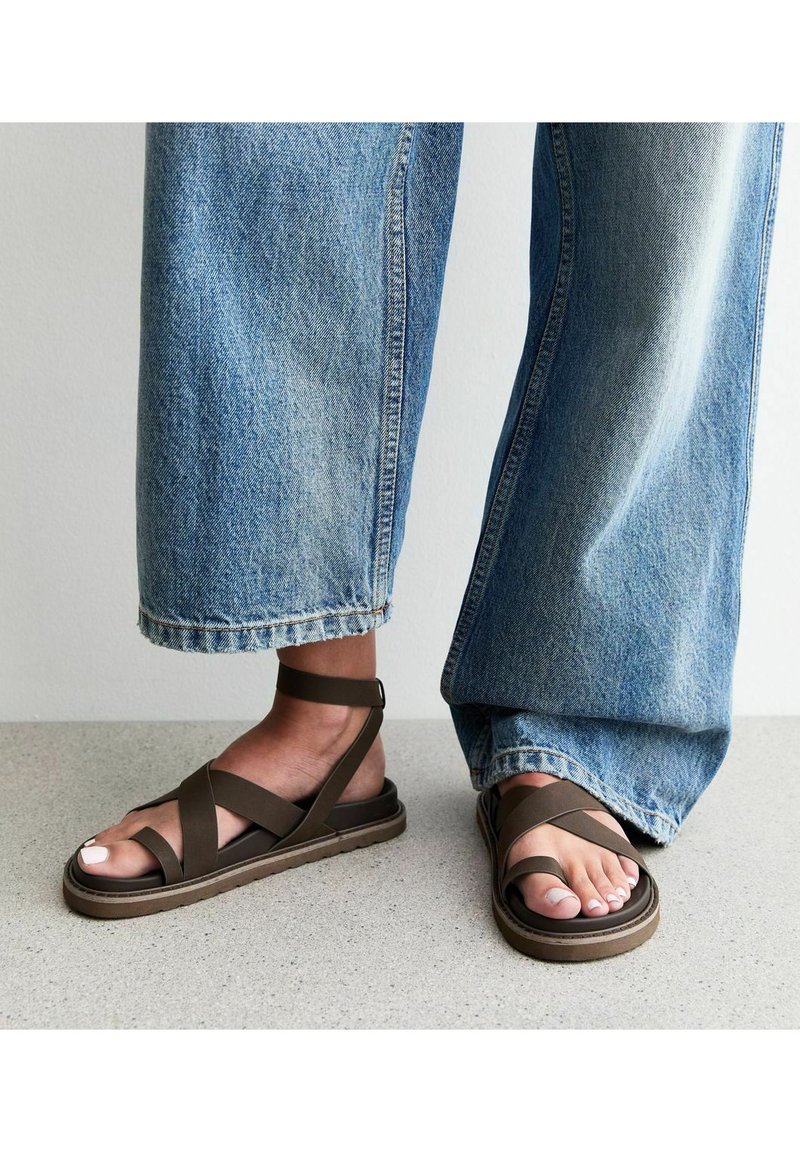 Sandali piatti marroni con più cinturini incrociati, cinturino alla caviglia e suola in gomma, abbinati a jeans a gamba larga. Le scarpe presentano una texture liscia.