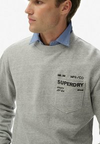 Hombre con sudadera gris y camisa azul de cuello por debajo, con "SUPERDRY" y otro texto impreso encima del bolsillo del pecho.