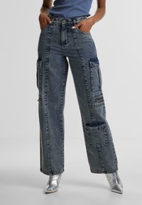 Denim cargo pants in dark wash with large side pockets, a high waist, and a straight leg fit, featuring subtle fading and a textured finish.