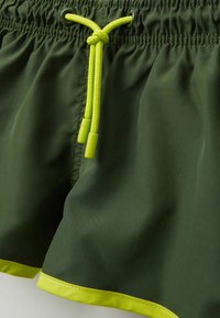 Zelené plavky s hladkou texturou, které mají jasně žlutou šňůrku na stažení a kontrastní žluté lemování.