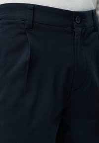 Κοντινό σε μέση σκούρου ναυτικού παντελονιού με κουμπί και θηλιές για ζώνη, φορεμένο με λευκό πουκάμισο που είναι μπα tucked.