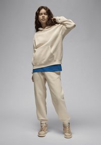 Felpa beige con tasca frontale, camicia blu a strati sotto, pantaloni sportivi beige abbinati e sneakers beige con accenti marroni. Design semplice.