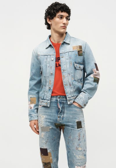 Νέος άντρας που φορά ανοιχτό μπλε τζιν μπουφάν και τζιν με μπαλώματα από ανάμεικτα υφάσματα, σε συνδυασμό με κόκκινο T-shirt με γραφικά, κοιτάζει στο πλάι.