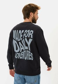 Zwarte fleece sweater met oversized grijze tekst op de rug met de tekst "MADE FOR DAILY ADVENTURES." Geribbelde manchetten en zoom.