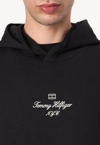Crna dukserica od mekog materijala s prednjim kengur džepom, kapuljačom s vezicom i izvezenim bijelim logotipom "Tommy Hilfiger NYC".