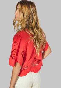 Cropped blouse rossa con bordi a ondina e intricati disegni intagliati. Tessuto leggero, maniche corte e vestibilità ampia.