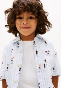 Νέο παιδί με σγουρά μαλλιά που φοράει μπλε ανοιχτό και λευκή ριγωτή μπλούζα με μικρές εικόνες παικτών μπάσκετ πάνω από λευκό μπλουζάκι.