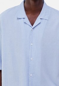 Světle modrá košile s krátkým rukávem a bílými vertikálními pruhy, s klopovým límcem a pěti knoflíky vpředu.