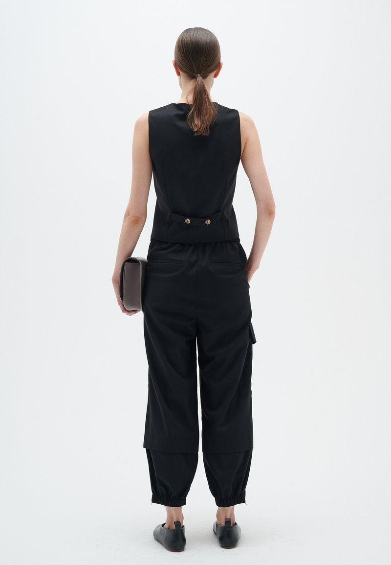 Frau mit braunem Pferdeschwanz, die ein ärmelloses schwarzes Oberteil und lockere schwarze Hosen trägt, hält eine graue Clutch und steht vor einem weißen Hintergrund.