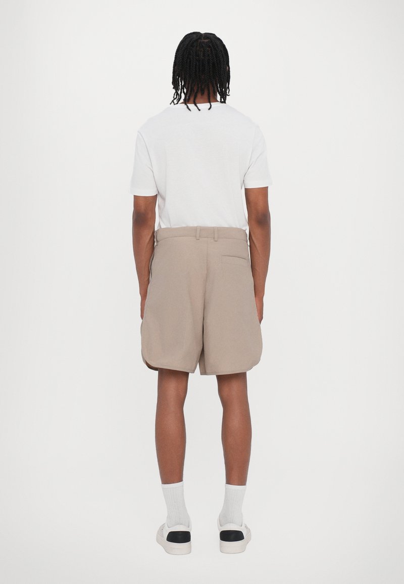 Beige shorts with a relaxed fit, side pockets, and a back pocket, paired with a white T-shirt and white socks with black detail.