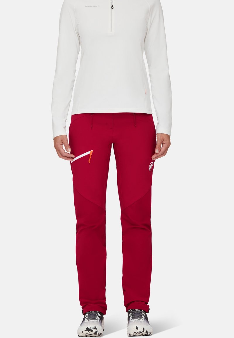 Personne portant un haut blanc à manches longues et un pantalon d'extérieur rouge avec des poches zippées, debout, chaussures blanches et grises visibles.