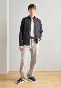 Camisa de botão às riscas pretas e brancas, jeans cinzentos, ténis pretos com solas brancas, de pé sobre um chão de madeira, roupa casual apresentada.