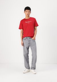 Camiseta de algodón roja con logo blanco y jeans de mezclilla gris. El modelo lleva zapatillas blancas, de pie sobre un fondo blanco liso.
