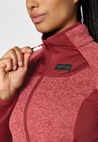 Giacca zip-up rossa con tessuto testurizzato, colletto alto e una toppa nera con logo sul lato sinistro del petto. Regolando la cerniera con una mano.