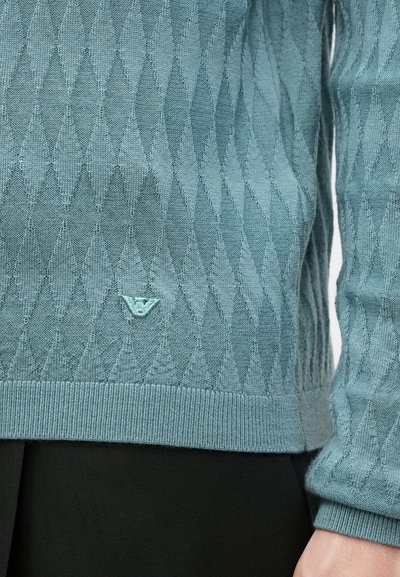 Ανοιχτό πράσινο πλεκτό πουλόβερ με μοτίβο ρόμβου, ριμπ μανσέτες και τελειώματα, με μια μικρή ετικέτα λογότυπου κοντά στο κάτω μέρος.