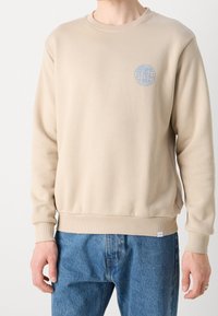 Felpa beige a girocollo con logo di un globo azzurro sul petto, indossata con jeans di denim blu, mostrando la parte superiore del corpo e le mani.
