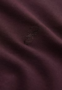 Burgundin värinen kangas, jossa on tekstuuri, ja jossa on kohokuvioitu hirviloogo. Materiaali näyttää hienovaraisia sävy- ja syvyysvaihteluja.