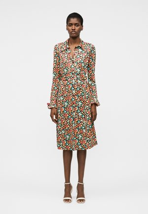 Γυναίκα που στέκεται φορώντας φόρεμα μακρυμάνικο, με γιακά, μήκους μέχρι το γόνατο με πολύχρωμο λουλουδάτο σχέδιο και λευκές γόβες με λουράκι στον αστράγαλο.