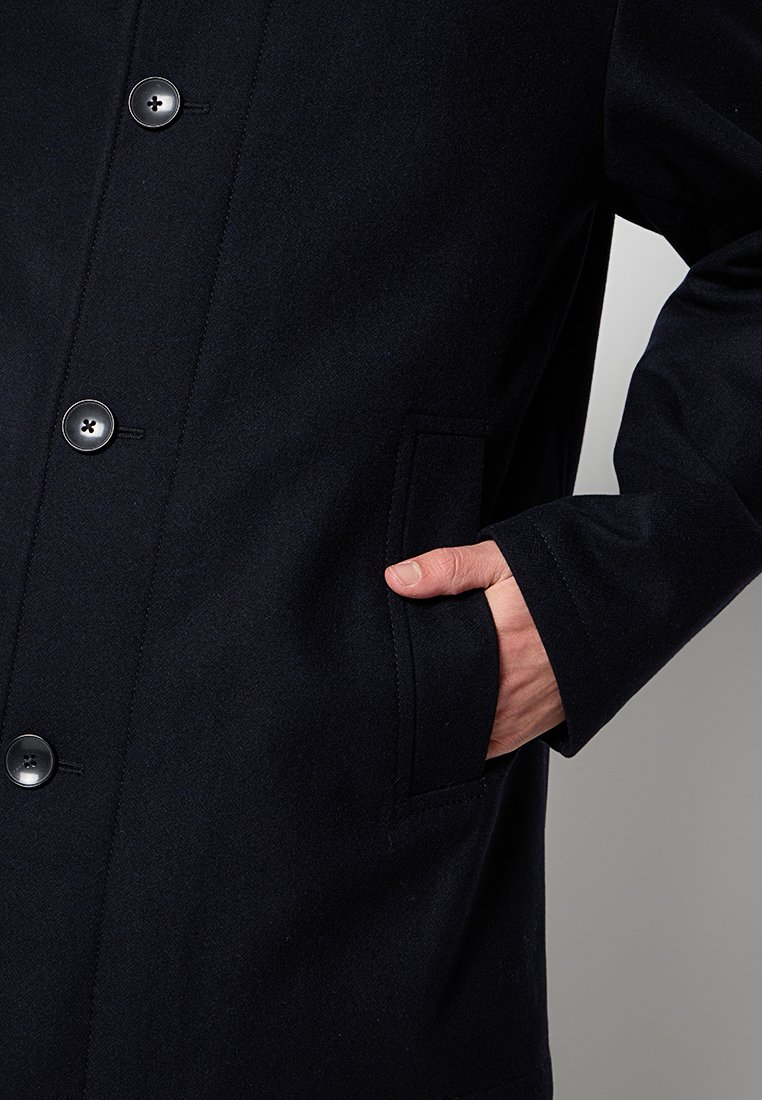 Cappotto nero con una texture liscia simile alla lana, design doppiopetto, sei bottoni e tasca laterale con una mano che riposa all'interno.