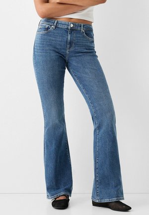 Person wearing high-waist blue flare jeans with front pockets, paired with black mesh shoes and a cropped white top, standing with arms crossed.