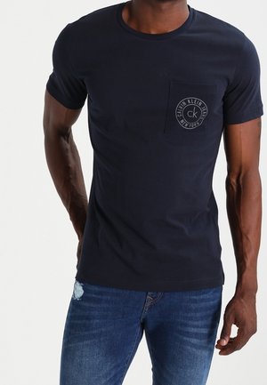 Homme portant un t-shirt noir Calvin Klein à manches courtes avec une poche logo et un jean bleu délavé, debout devant un fond blanc.
