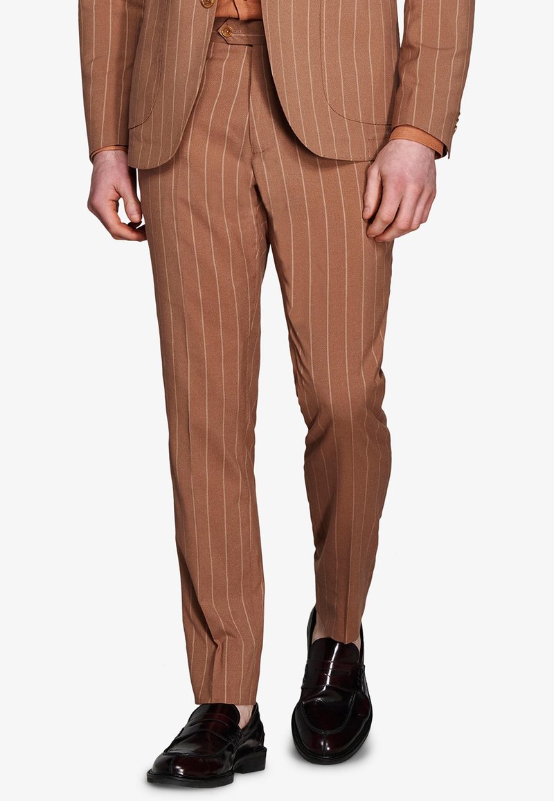 Mężczyzna ubrany w brązowe spodnie w prążki od garnituru z dopasowaną marynarką oraz czarne eleganckie buty, stojący na białym tle.