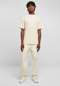 Tricou din bumbac bej, cu mâneci scurte, asociat cu pantaloni bej deschis asortați și adidași albi; design minimal, cu o textură ușor vizibilă.