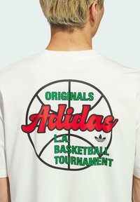 Bijela pamučna majica s velikim grafikama na leđima, s crvenim natpisom "Adidas", zelenim natpisom "L.A. BASKETBALL TOURNAMENT" i konturama košarkaške lopte.
