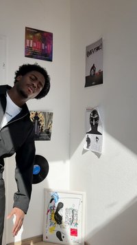 Una figura con giacca nera si appoggia a un muro decorato con poster musicali, un disco in vinile e un'opera d'arte astratta con colori vivaci.