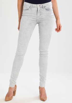 Person iført lysegrå skinny jeans og brune højhælede sko, stående med benene let spredt mod en ensfarvet baggrund.