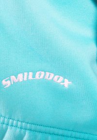 Tissu bleu clair avec texte brodé blanc "SMILODOX" en forme d'arc au-dessus d'une couture cousue.