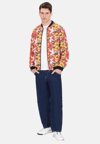 Giacca bomber rossa con motivo floreale giallo, bordi neri, t-shirt bianca, jeans blu scuro e sneakers bianche. Stile casual, vestibilità rilassata.