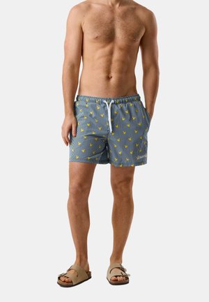 Oberkörperfreier Mann trägt blau-weiß gestreifte Badeshorts mit gelben Entenprints und beige Sandalen vor einem schlichten Hintergrund.