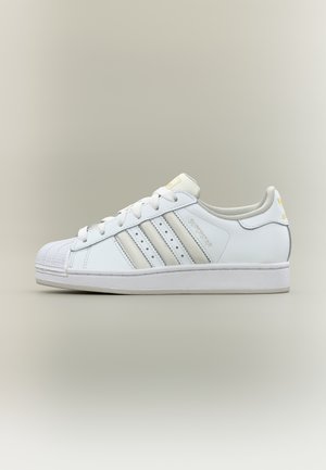 Baskets Adidas Superstar blanches avec trois bandes grises, bout renforcé en forme de coquille, et inscription "Superstar" sur le côté, montrées de profil sur un fond clair.