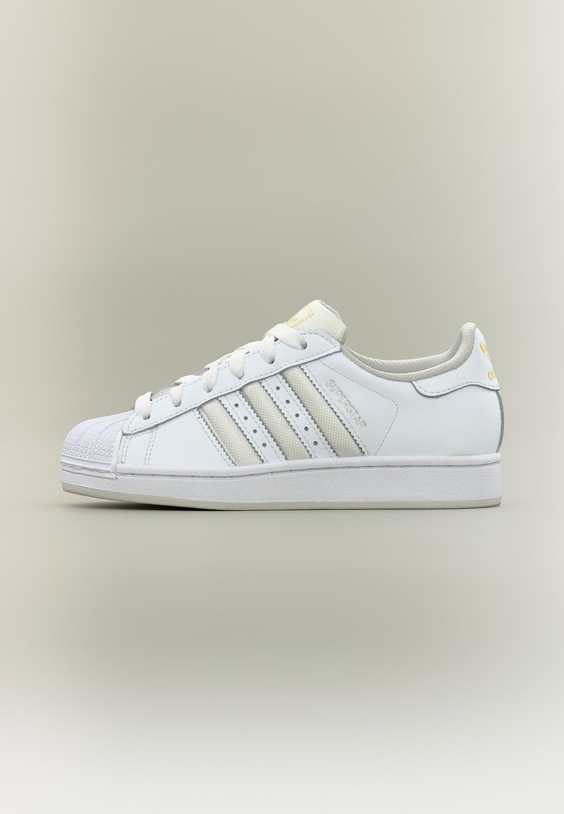 Λευκό αθλητικό παπούτσι Adidas Superstar με τρεις γκρι ρίγες, προστατευτικό καβουκιού και το κείμενο "Superstar" στο πλάι, εμφανιζόμενο σε προφίλ σε φωτεινό φόντο.