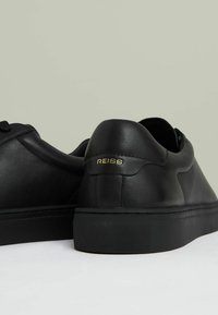 Zapatillas de cuero negro con un acabado suave, que presentan una suela texturizada y el logo dorado "REISS" en el talón. Diseño minimalista, forma redonda.