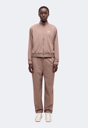 Personne portant un survêtement mauve zippé avec des poignets élastiques et des baskets blanches, debout face à un fond uni.