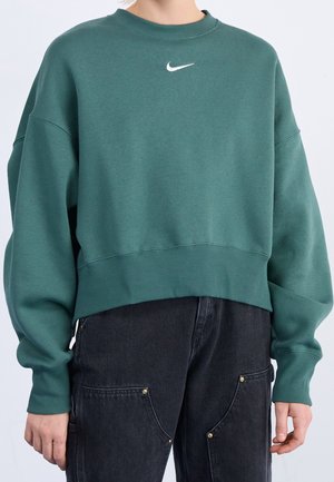 Person wearing a green cropped Nike sweatshirt and black high-waist jeans with large front pockets against a plain background.