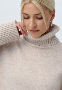 Μπεζ πουλόβερ με ζιβάγκο, με ριχτές μανσέτες και γιακά. Μαλακή πλεκτή υφή με διακριτικές λεπτομέρειες και ένα μικρό κεντημένο λογότυπο στη μέση.