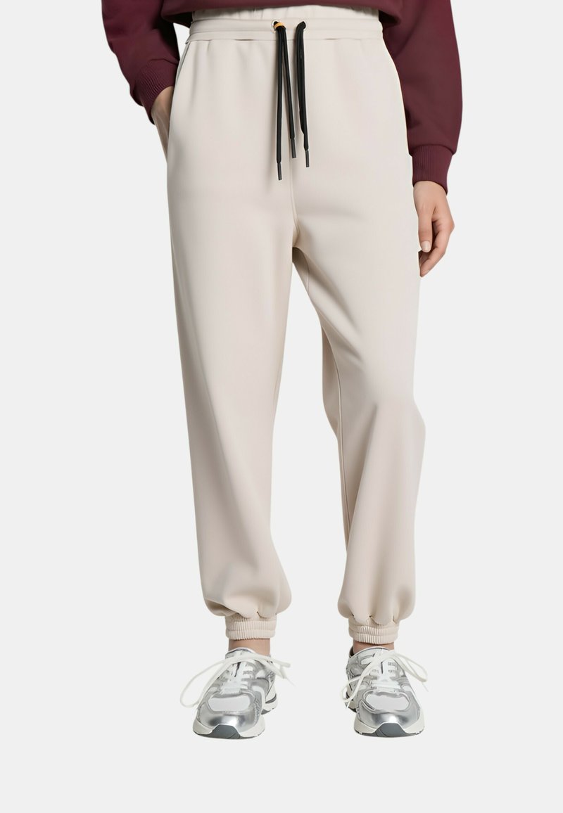 Pantaloni jogger beige con polsini elastici e coulisse nere, indossati con sneakers argento e bianche, mostrati dalla vita ai piedi.
