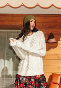 Pull blanc en maille torsadée avec des manches surdimensionnées, superposé à une jupe à volants à motif floral rouge et vert. Bonnet côtelé vert olive.