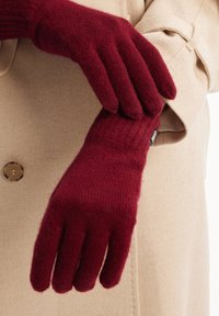 Burgundy stickade handskar med ribbat mönster, visade mot en ljus beige kappa. Har en etikett vid handleden och en åtsittande design.