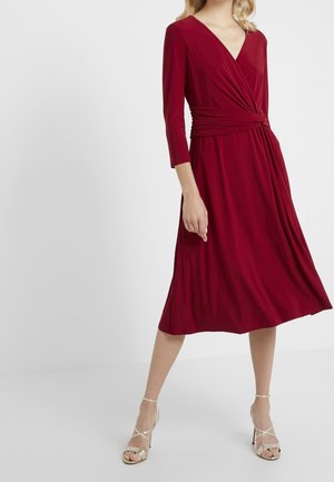 Femme portant une robe bordeaux à manches longues et longueur genou avec une taille nouée, ainsi que des sandales à talons hauts beiges, debout sur un fond blanc.