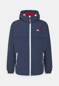 Mörkblå vindjacka med huva, vit dragkedja och sidofickor; har en röd innerfoder och en logotyp på bröstet.