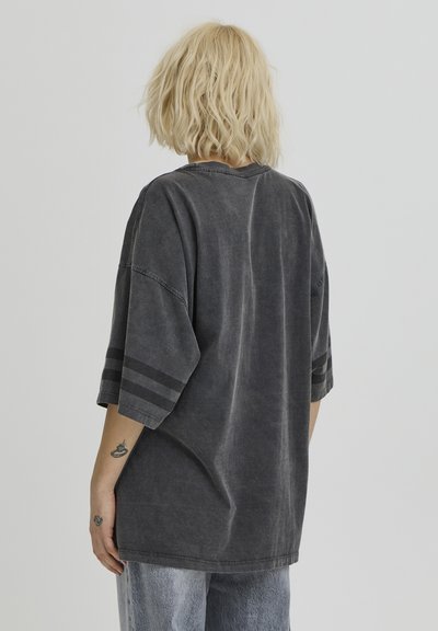 T-shirt gris surdimensionné en tissu doux, avec manches courtes ornées de deux rayures horizontales et un col rond. Texture vintage subtile.