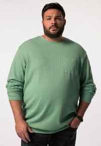 Camisa verde de manga larga con tejido texturizado, cuello redondo y un bolsillo en el pecho. Combinada con pantalones vaqueros oscuros.