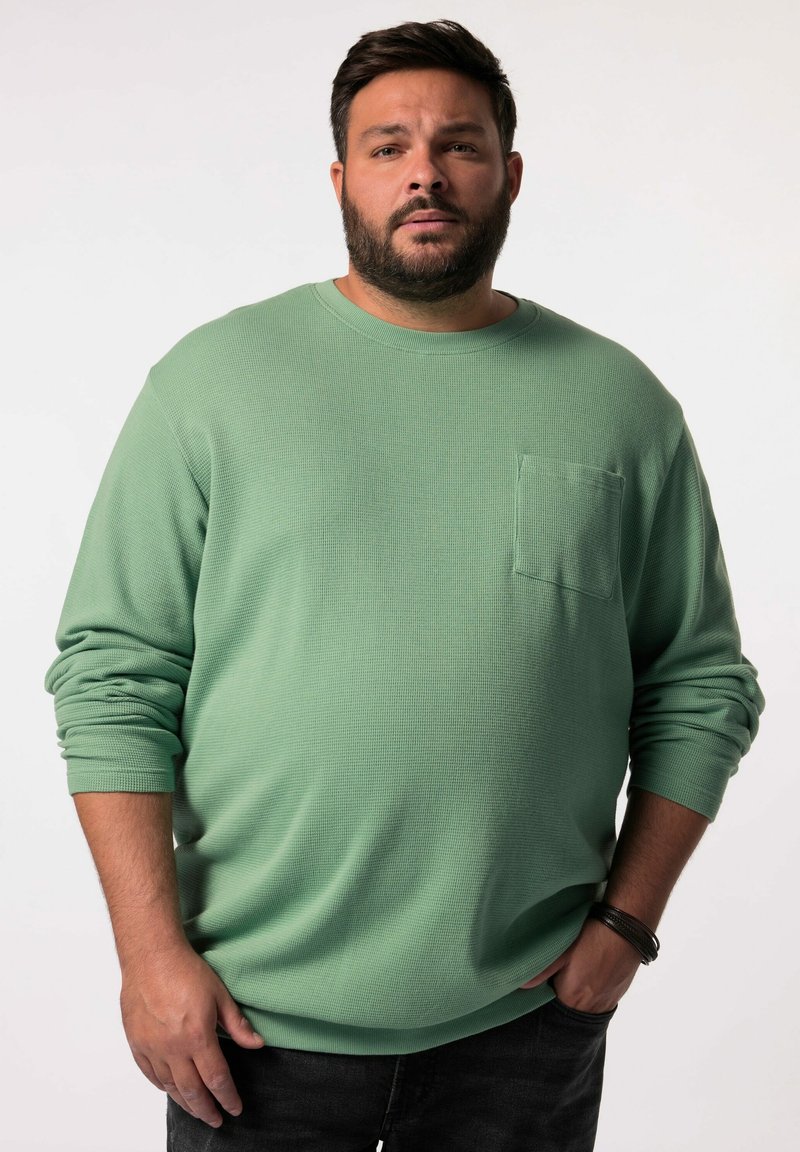 Camisa verde de manga larga con tejido texturizado, cuello redondo y un bolsillo en el pecho. Combinada con pantalones vaqueros oscuros.