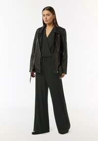 Black leather jacket over a green jumpsuit with wide legs, drawstring waist, and subtle shimmer. Model stands against a white background.