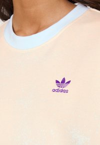 Felpa di colore pesco chiaro con una consistenza morbida, dotata di un colletto a girocollo blu a costine e un logo Adidas ricamato di colore viola sul petto.