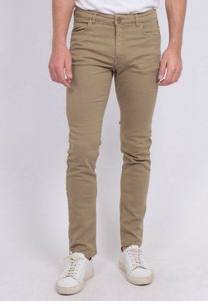 Homme portant un pantalon beige slim et des baskets blanches, debout devant un fond clair uni.